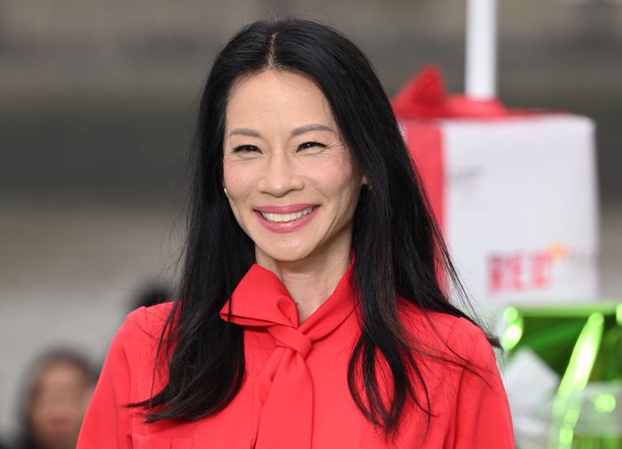 Lucy Liu beaming with a smile, dressed in a chic blouse with a bow tie detail, standing outdoors with a soft, blurred backdrop.