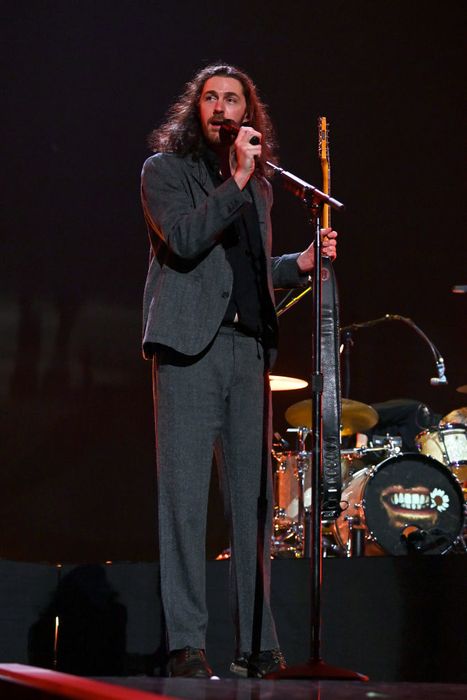 Hozier, dressed in a dark suit, performs on stage, speaking into a microphone while holding a guitar, with drums visible in the background.
