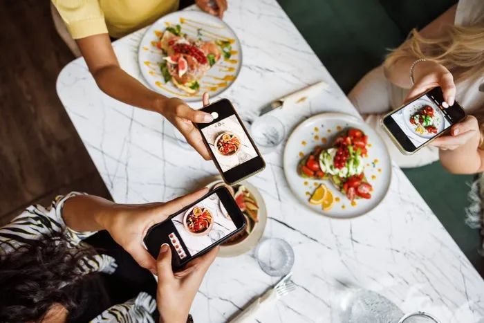 A group of three individuals snaps pictures of beautifully arranged dishes at a restaurant, capturing their meals on their smartphones.