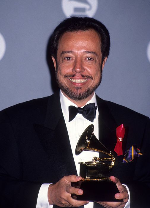 A person in a tuxedo smiles at the camera while holding a Grammy award.