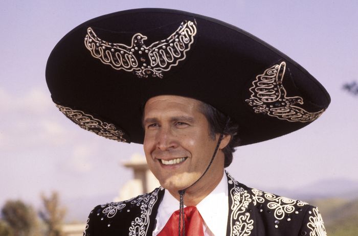 A person wearing an elaborate sombrero and a matching embroidered suit smiles cheerfully while standing outdoors.