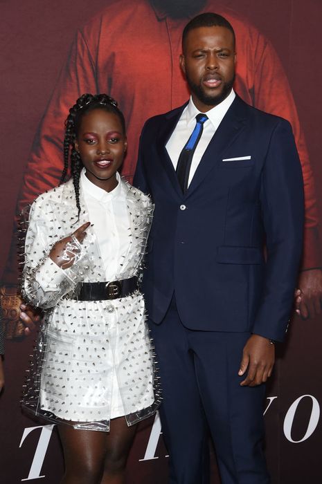 Lupita and Winston strike a pose together; she stuns in a studded sheer jacket over a light ensemble, while he looks sharp in a suit with a shirt and tie. She stands just above his shoulder.