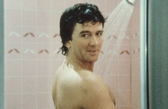 A person in a shower, glancing back with a playful expression, with a pink-tiled wall visible in the background.