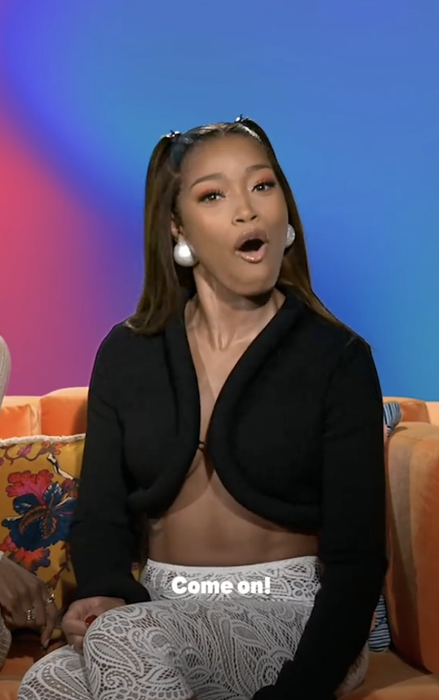 A person speaking passionately, wearing a black top with striking earrings, seated on a vibrant couch. Subtitles on the image say: "Come on!"