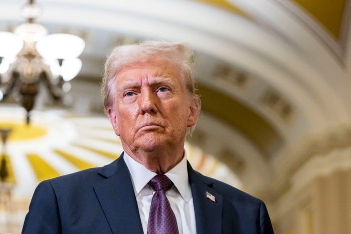 Donald Trump, wearing a suit, stands with a serious expression in a beautifully decorated hallway.