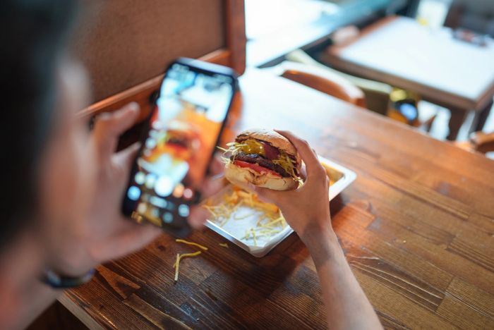 A person snapping a photo of a gourmet burger with a smartphone at a wooden table, capturing the culinary details for social media