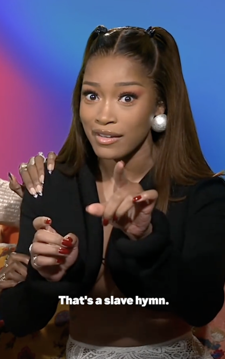 A person making a gesture while speaking, wearing a chic black top. Text on image: "That's a slave hymn."
