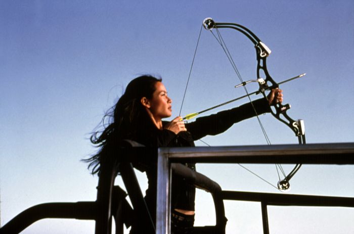 Lucy Liu practicing archery with a modern bow, aiming at a target on an outdoor platform.