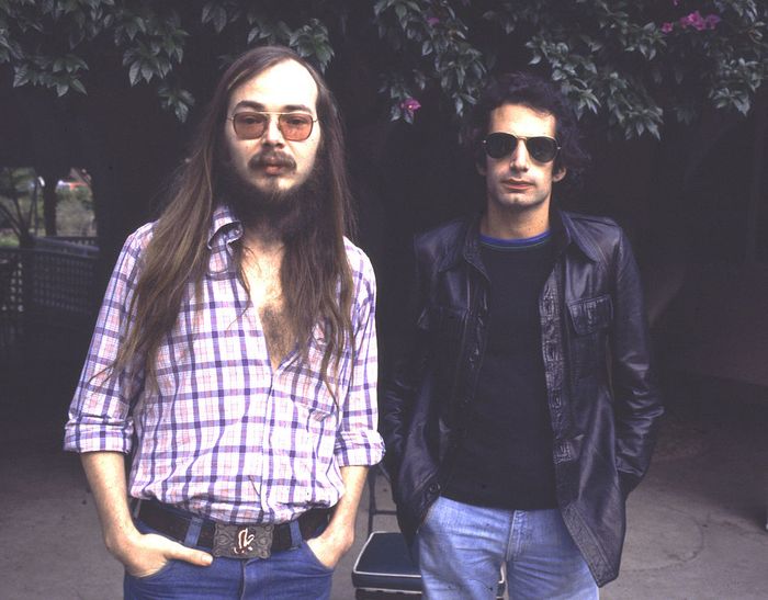 Two men with long hair and sunglasses stand outdoors. One wears a plaid shirt and jeans, while the other sports a black leather jacket and jeans, with trees in the background.