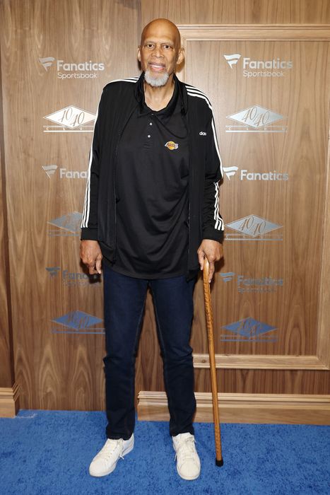 Kareem appears on the red carpet wearing a casual jacket, dark shirt, and jeans, while holding a cane.