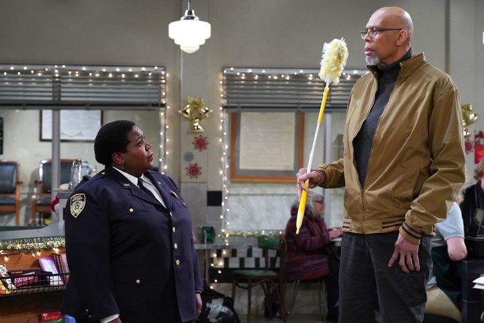 Lacretta and Kareem share a comedic moment: she’s in a police uniform, looking up at him as he holds a mop while dressed in a casual jacket. She doesn’t quite reach his chest.