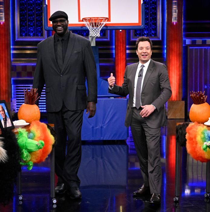 Shaq and Jimmy, both suited up, share a laugh on a talk show set adorned with basketball-themed decorations. Jimmy barely reaches Shaq’s shoulder.