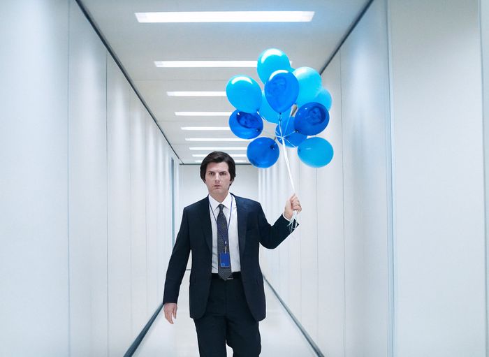 A person in a suit walks down a hallway, holding a bunch of blue balloons.