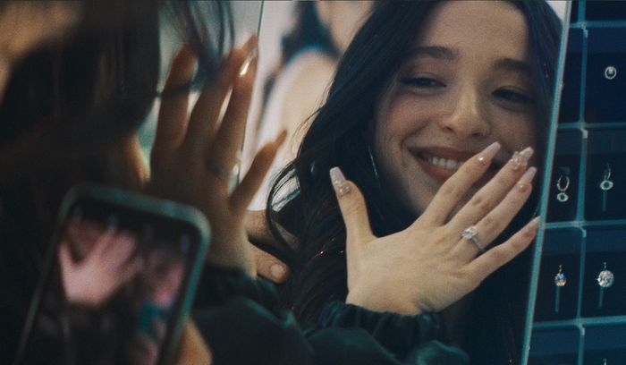 A woman smiles, showcasing a ring on her hand in a jewelry store, her reflection visible in a mirror.