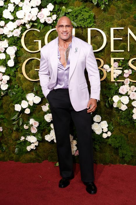 The Rock grins on the red carpet, dressed in a pastel blazer, silk shirt, and dark trousers, with a floral backdrop and the