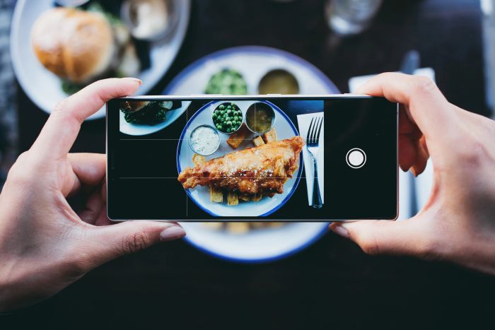 A person capturing a photo of a meal featuring fish and chips, peas, and sauce