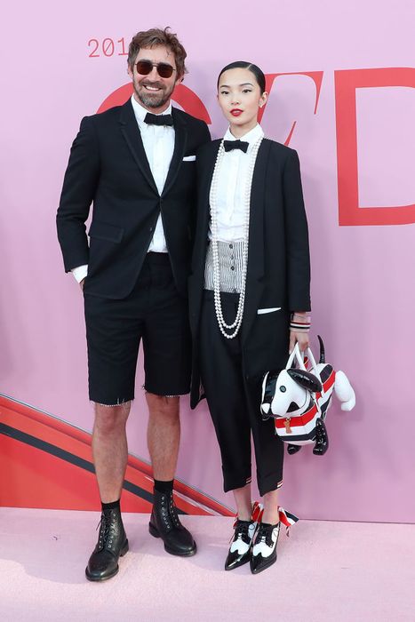 Lee Pace and Xiao Wen Ju grace the red carpet; he sports a tuxedo jacket paired with shorts and boots, while she stuns in a tuxedo with pearl accessories, holding a dog-themed bag. In heels, she reaches his eye level.
