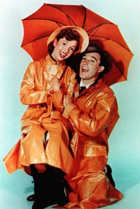 Debbie and Gene, both wearing raincoats, smile while posing with an umbrella.