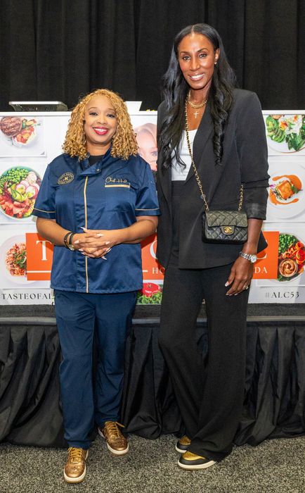 Huda and Lisa pose in front of a culinary-themed backdrop: Huda wears a professional chef’s uniform, while Lisa stuns in an elegant blazer and pants. Huda reaches just up to Lisa’s shoulder.