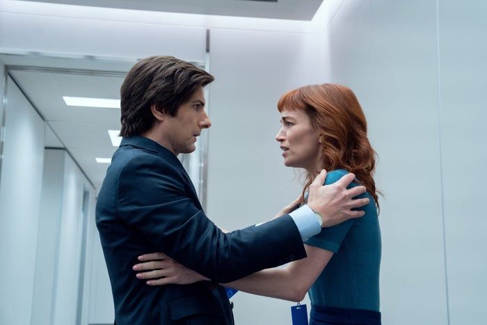 Two characters from the show stand facing each other, engaged in a tense conversation in a modern, white hallway.