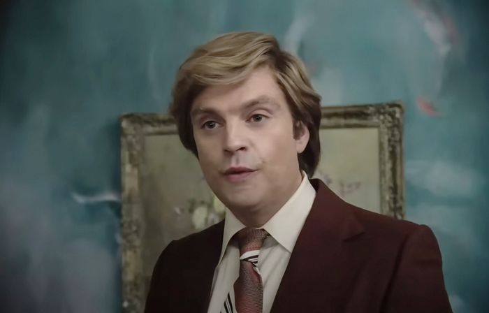 Sebastian Stan portrays Donald Trump in a suit and tie, standing in front of a framed painting, with a softly blurred background.