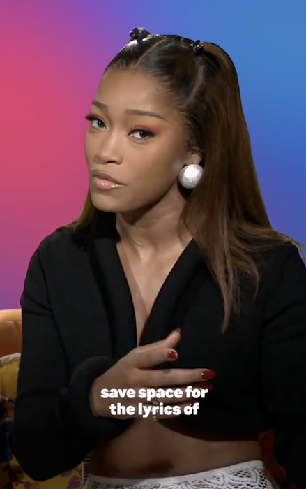 A person with long hair, wearing a black top and big earrings, speaks; the text says "save space for the lyrics of"