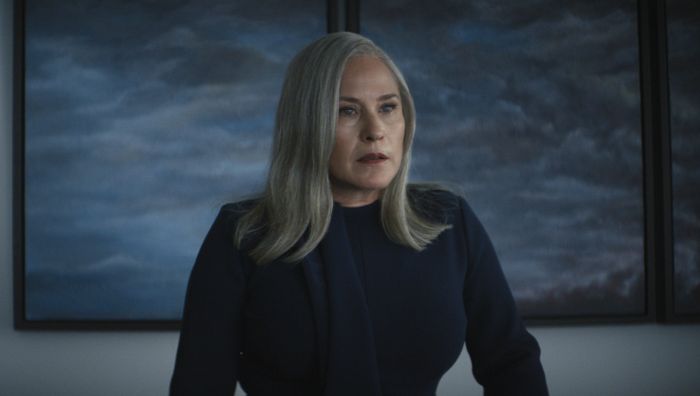 Harmony, with long hair, stands in front of abstract artwork, dressed in a formal dark outfit.