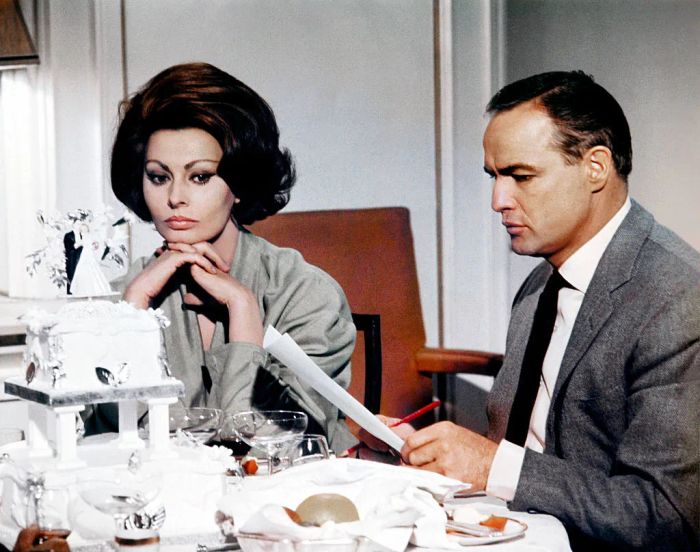 Sophia and Marlon sit at a table with a wedding cake in front of them. Sophia, dressed in a vintage gown, gazes thoughtfully into the distance while Marlon, in a suit, reads a newspaper.