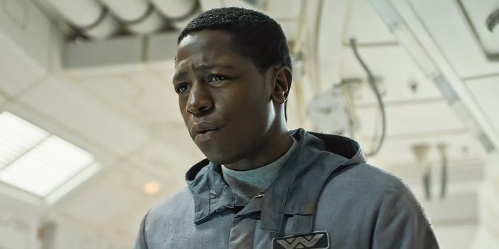A person in a futuristic environment, donning a gray jacket with a logo, gazes ahead with a determined look.