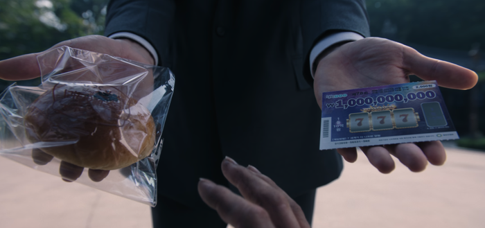 A close-up of two hands; one holds a plastic-wrapped bun, the other a scratch-off ticket revealing three sevens, implying a link between the two.