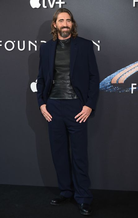 Lee, dressed in a sleek dark suit and leather shirt, poses on the red carpet against an Apple TV+ event backdrop.