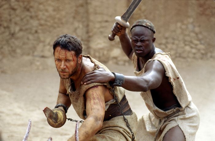Djimon Hounsou and Russell Crowe in Gladiator, one wielding a sword while defending the other. They appear intense and battle-worn.