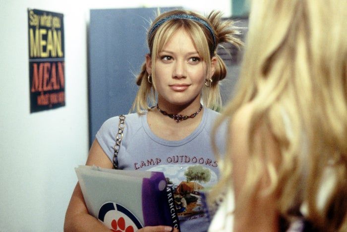 A person dressed casually in a graphic tee and headband, clutching a folder, stares at another individual. A poster reads, 