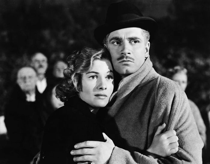 A memorable film moment where Joan and Laurence share an embrace; she appears worried while he, dressed in a coat and hat, seems to be offering protection.
