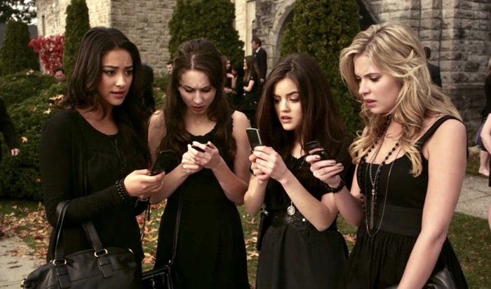 Four young women in elegant black dresses stand closely together, anxiously checking their phones with concerned expressions.