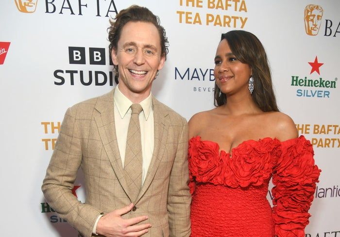 Tom and Zawe at the BAFTA Tea Party. He dons a patterned suit and tie, while she wears an elegant off-shoulder dress adorned with floral details.