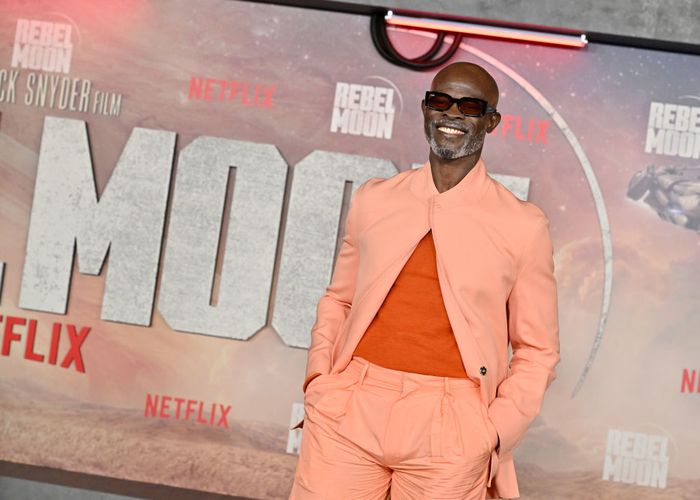 A person dressed in a stylish tan suit and sunglasses at a Netflix event, with a large 