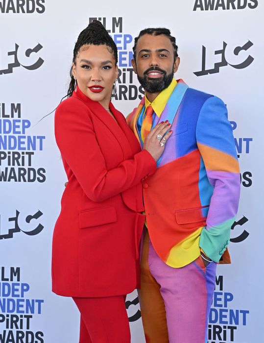 At an awards ceremony, Emmy strikes a bold look in a monochrome suit, while Daveed dazzles in a multicolored ensemble. The two smile as they pose together on the red carpet.