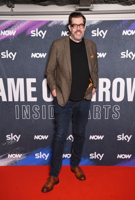 Richard smiles on the red carpet, dressed in a blazer, shirt, and jeans.