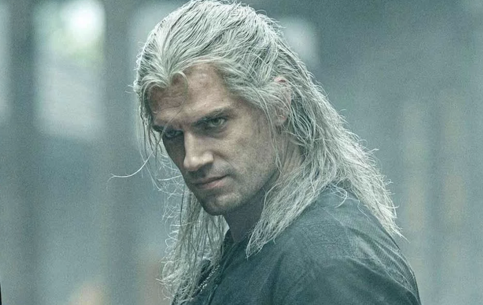 A man with long silver hair, dressed in a medieval-inspired leather outfit, gazes intently in a misty environment.