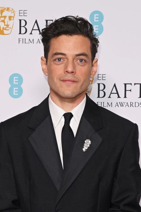 Rami Malek, in a black suit with a floral lapel pin, poses in front of a BAFTA event backdrop.