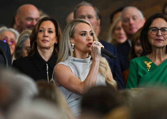 Carrie Underwood passionately performs into a microphone at a formal event, with Kamala Harris standing behind her.