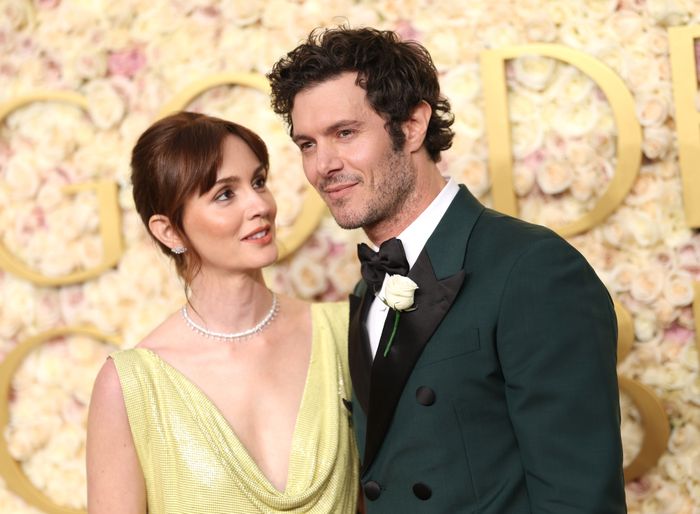 Adam, dressed in a tuxedo with a rose boutonniere, and Leighton, in a chic sleeveless gown, strike a pose on the floral-themed red carpet, radiating elegance.
