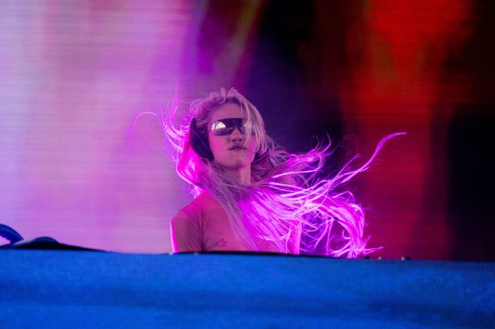 A DJ, wearing sunglasses, energetically performs on stage with long hair flowing, bathed in vibrant stage lighting.