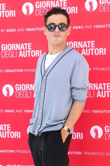 Rami Malek, wearing a casual cardigan and sunglasses, poses against a backdrop featuring the words 