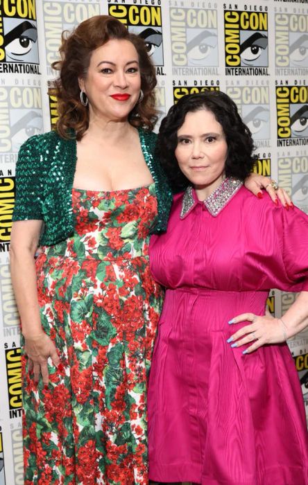 At Comic-Con, Jennifer wears a floral dress with a sparkly jacket, while Alex sports a bright cinched dress with a detailed neckline. Alex reaches up to Jennifer's earlobe.