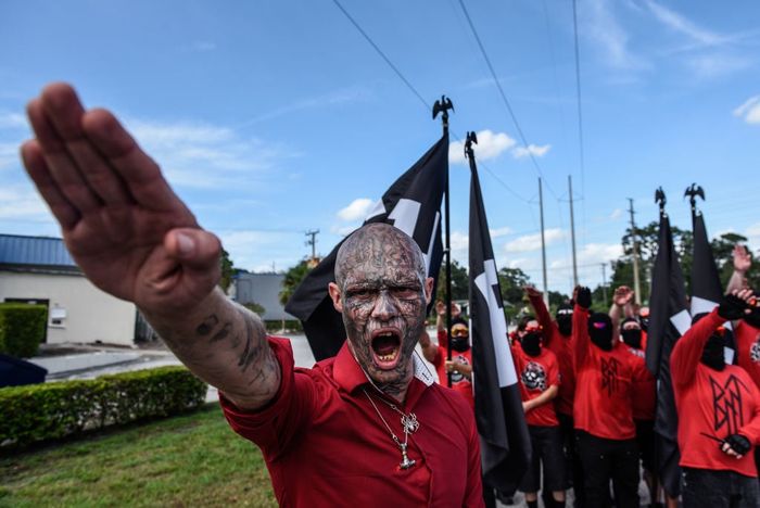 A group of individuals in red shirts making controversial hand gestures during a protest, with flags visible in the background.