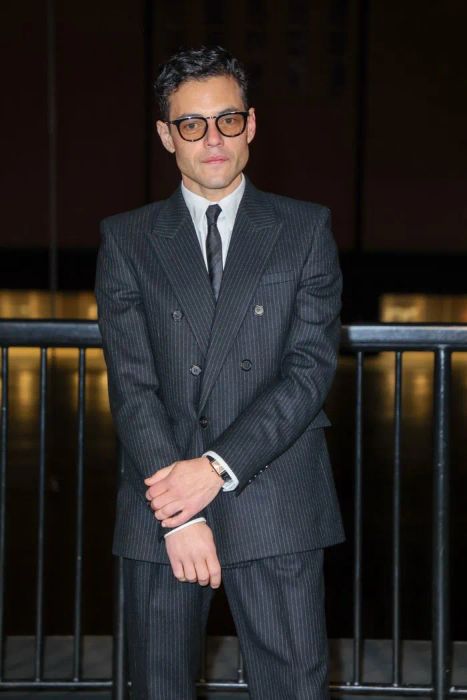 Rami Malek, dressed in a pinstripe suit and glasses, standing with arms crossed indoors.
