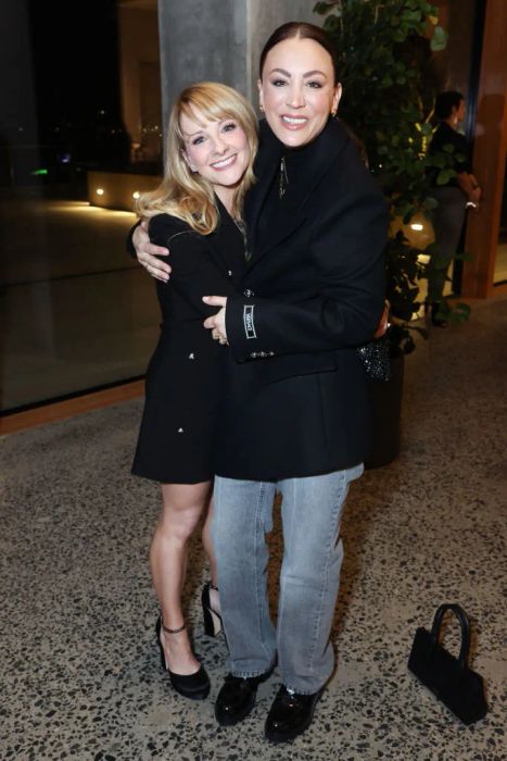 Melissa and Kaley share a hug, grinning at an event. Melissa is dressed in a dress and heels, while Kaley wears a blazer and jeans. A small handbag sits on the floor. Melissa's height reaches just to Kaley's eyes.