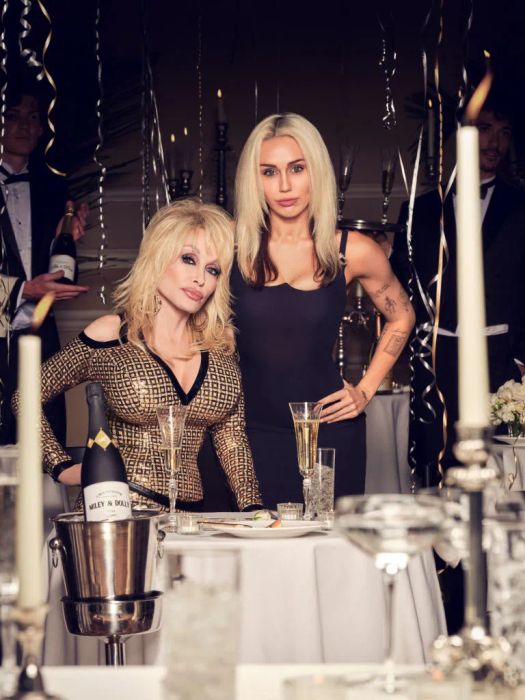 At a formal gathering, Dolly is dressed in a patterned cold-shoulder top, while Miley wears a sleeveless dress. They stand amid party decorations. Dolly reaches up to Miley's shoulder.